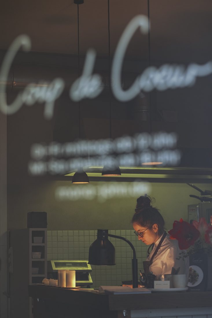 Blick durch das Schaufenster mit „Coup de Cœur“-Schriftzug auf Mona bei der Arbeit